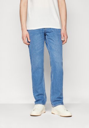 Persoon die lichtblauwe rechte jeans, witte sneakers en een wit T-shirt draagt, staand tegen een effen lichte achtergrond.