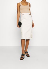 Beige fitted tank top with square neckline, paired with a white knee-length skirt featuring a side slit. Model holds a black clutch and wears black mules.