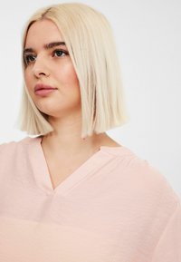 Vaaleanpunainen paita, jossa on V-pääntie ja pehmeä kangas; sisältää väljyyttä ja lyhyet hihat, korostaen minimalistista tyyliä.