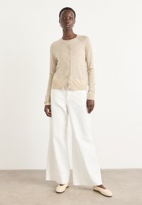 Beige cardigan med rund halsringning, knäppning med knappar och ribbade ärmslut, kombinerad med vida vita byxor och ljusa skor. Slät textur.