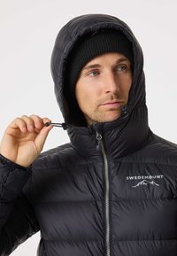 Svart puffjacka med huva, med horisontellt quiltade sektioner, dragkedja fram och justerbara snoddar. Logotyp tryckt på bröstet.