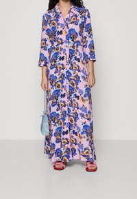 Kwiatowa sukienka maxi w kolorach niebieskim, pomarańczowym i różowym, z guzikami z przodu i rękawami do trzech czwartych. Połączona z różowymi sandałami i jasnoniebieską torbą.