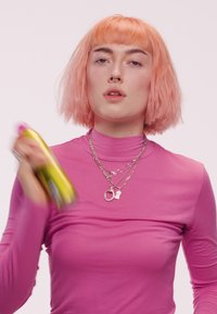 Personne avec des cheveux courts pêche portant un col roulé rose et des colliers en argent superposés, secouant un spray doré et rose.