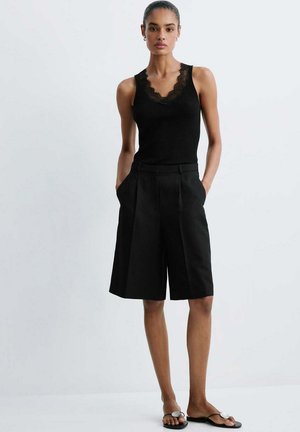 Femme debout les mains dans les poches, portant un débardeur noir avec bordure en dentelle, un short noir longueur genou et des sandales noires avec des accents blancs.