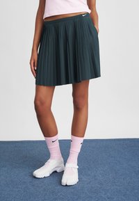 Nokrāsota tumši zaļa tenisa svārku ar krokām, ar elastīgu jostu, apvienota ar rozā zeķēm un baltām slip-on kurpēm. Fons ir pelēks un zils paklājs.