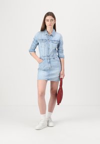 Denim obleka v svetlo modri barvi z dolgimi rokavi, gumbi spredaj, dvema žepoma na prsih in oprijetim pasom. V kombinaciji z belimi supergami in rdečo torbico.