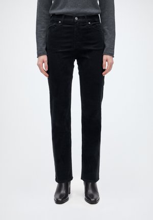 Modèle portant un jean noir à coupe droite, un haut gris et des bottines noires à talons, sur un fond clair uni.