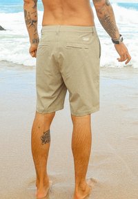 Shorts de bain beiges, de longueur mi-cuisse, avec poches arrière et une petite étiquette de logo. Tissu doux, portés sur une plage de sable près des vagues de l'océan.