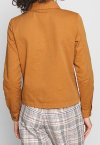 Světle hnědá bavlněná bunda s límcem, knoflíkovými manžetami a rovnými lemy, doplněná kostkovanými kalhotami s pruhy v červené, černé a žluté barvě.