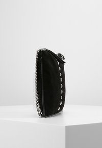 Clutch de ante negra con cadena de plata y tachuelas en forma de pirámide, de pie sobre una superficie blanca contra un fondo liso.