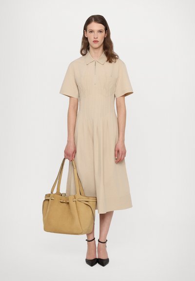 Beige lyhythihainen mekko, jossa on edessä vetoketju, pleat-tyksentelyä ja väljä malli. Mallilla on keltainen mokkanahkainen käsilaukku ja mustat korkokengät.