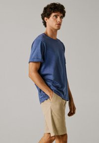 Käharate juustega mees sinise triibulise T-särgi ja beežide lühikeste pükstega, seisab käed taskutes tavalise tausta ees.