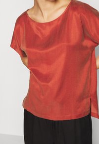 Kortærmet rød bluse med bred halsudskæring, glat tekstur og sideskår. Kombineret med sorte bukser, der fremhæver en afslappet pasform.