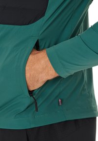 Il top a maniche lunghe di colore teal presenta un pannello nero sul petto e una tasca con zip sul lato, con una texture liscia e leggera.