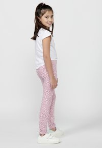 Enfant portant un t-shirt blanc à manches courtes et un legging en imprimé léopard rose, associé à des baskets blanches avec des bandes Velcro.