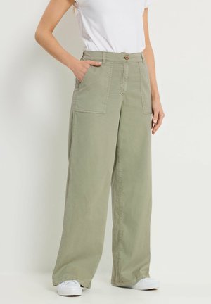 Femme portant un pantalon larges jambes de couleur olive clair avec des poches plaquées à l'avant, une fermeture à boutons, des baskets blanches et un haut à manches courtes blanc.