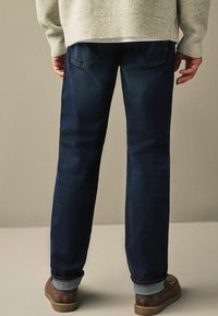 Donkerblauwe denim jeans met een rechte snit, zichtbare achterzakken en opgerolde mouwen. Draag met bruine instap schoenen.