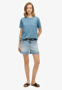 Blauwe katoenen t-shirt met een borstzak en logo, gecombineerd met versleten denim shorts en zwarte slipper sandalen. Casual zomeroutfit.