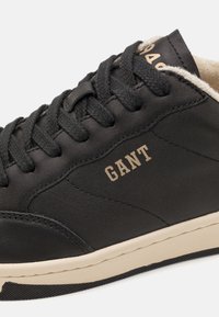 Schwarze Ledersneaker mit runder Zehenpartie, strukturiertem Obermaterial und kontrastierender beiger Sohle. Verfügt über ein goldfarbenes Logo an der Seite und flache schwarze Schnürsenkel.