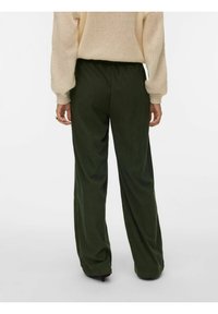 Osoba oblečená v širokých tmavě zelených kalhotách a béžovém pleteném svetru, stojící zády ke kameře.