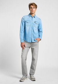 Camisa de mezclilla azul claro con botones a presión, yokes puntiagudos y dos bolsillos en el pecho, combinada con pantalones grises claros y zapatillas grises.