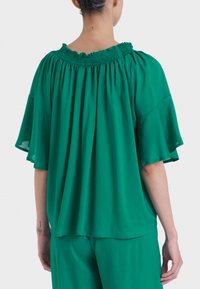 Top verde, largo e morbido con scollatura arricciata e maniche corte a volant. Realizzato in tessuto leggero, presenta una silhouette fluida e informale.