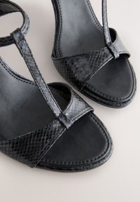 Sandale negre cu toc, cu un model texturat de piele de șarpe. Design cu vârf deschis, barete subțiri și talpă din piele netedă.