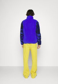 Chaqueta de forro polar en azul vibrante con una manga negra estampada, combinada con pantalones de pierna ancha amarillos brillantes. Diseño sencillo y textura suave.