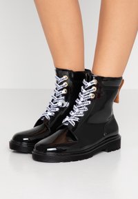 Bottines en cuir verni noir avec des lacets blancs aux marques, œillets dorés et une semelle texturée. Comprend un tirant de talon marron pour un enfilage facile.