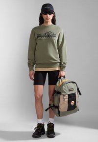 Felpa verde con logo, pantaloni corti neri e stivali neri. Porta uno zaino beige e verde con tasca in rete e dettagli arancioni.