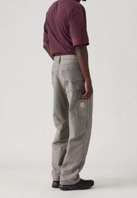 Pantalones cargo de mezclilla gris con costuras visibles, bolsillos laterales y una etiqueta en el bolsillo trasero. Llevados con una camiseta de manga corta burdeos y zapatos negros.