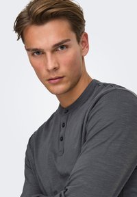 Donkergrijze henley met lange mouwen, voorzien van een ronde hals, drie zwarte knopen en een zachte, gestructureerde stof. Model met een neutrale uitdrukking.