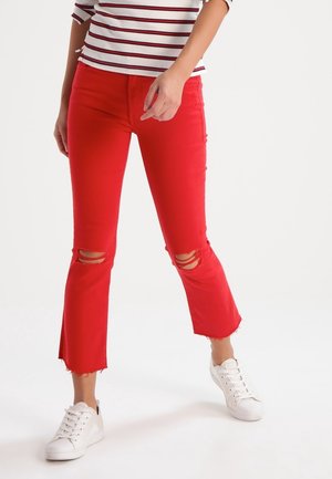 Kobieta w czerwonych podartych krótkich spodniach, białych trampkach i białej koszulce z długim rękawem z czerwonymi paskami na jednolitym tle.