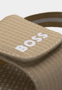 Κοντινό πλάνο μπεζ λουριού σανδάλου με ανάγλυφο λευκό λογότυπο "BOSS" στην κορυφή και μοτίβο στο πέλμα από κάτω.