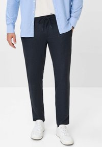 Dunkelgraue Hose mit Kordelzugbund, glatter Textur und schmal zulaufendem Bein, kombiniert mit weißen Sneakern und einem hellblauen Hemd mit Knöpfen.