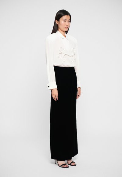 White blouse with oversized bow detail, paired with a floor-length black velvet skirt and black strappy heels. Smooth textures, minimalist design.