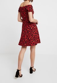 Žena v červených off-shoulder šatech s černým leopardím vzorem a černými blokovými podpatky s kotníkovými pásky, stojící na jednobarevném bílém pozadí.