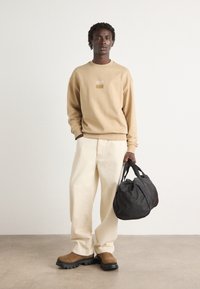 Beige sweatshirt with a square logo, cream wide-leg pants, brown boots, and a black soft bag with dual handles.