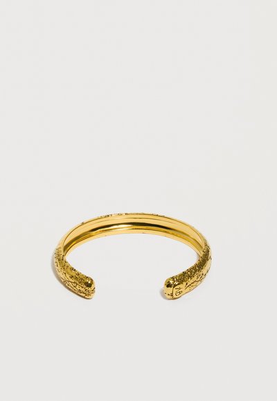 Λεπτό χρυσό βραχιόλι μανσέτα με υφασμένη σχεδίαση, ανοιχτό στο άκρο με διακριτικές χαραγμένες λεπτομέρειες πάνω σε μια επίπεδη λευκή επιφάνεια.