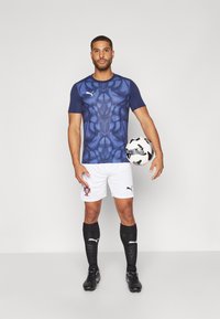 Mørkeblå kortermet sports skjorte med teksturert mønster, hvite shorts med logo, svarte sokker og svarte fotballsko, holder en fotball.