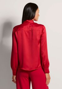Rote Satinbluse mit langen Ärmeln, hohem Kragen und Rückenschulternaht, samt glatter Textur und geknöpften Manschetten.