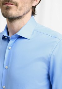 Camicia azzurro chiaro con bottoni, realizzata in tessuto liscio, con colletto a punta e una sottile lucentezza, che mostra una silhouette aderente e bottoni bianchi standard.