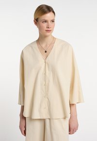 Cremfarbene, durchgeknöpfte Bluse mit V-Ausschnitt, weiten Ärmeln und einer weichen Textur. Die Vorderseite hat gleichmäßig verteilte Knöpfe in der Mitte.