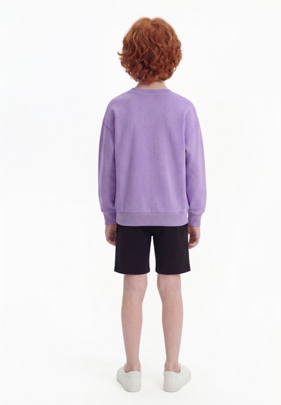 Child with curly red hair wearing a lavender sweatshirt, black shorts, and white shoes standing facing away on a white background.