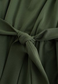 Grönt tyg med en vriden knutdetalj, slät textur och flödande fall, med subtila nyansvariationer över ytan.