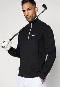 Μαύρος πλεκτός πόλο πουλόβερ γκολφ με ψηλό γιακά, μπροστινό φερμουάρ μέχρι τη μέση, ραβδωτές λεπτομέρειες στα μανίκια και ένα μικρό λευκό λογότυπο στο στήθος.