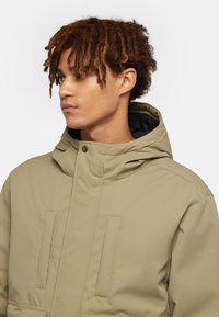 Beige huva jacka med två vertikala fickor, slät textur och knäppning med snodd. Inuti har den svart foder.