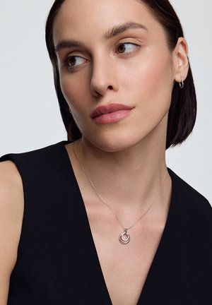 Srebrny naszyjnik z okrągłym wisiorkiem, który ma teksturowany zewnętrzny pierścień i gładki wewnętrzny pierścień, noszony na ciemnym topie.