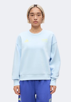 Personne aux cheveux blonds courts portant un sweat-shirt bleu clair et un pantalon bleu avec des logos jaunes Adidas, debout devant un fond uni.