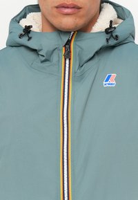 Lyseblå K-WAY-jakke med hette, med hvit glidelås og fargerike striper som detaljer, teksturert fôr og justerbare snorer.